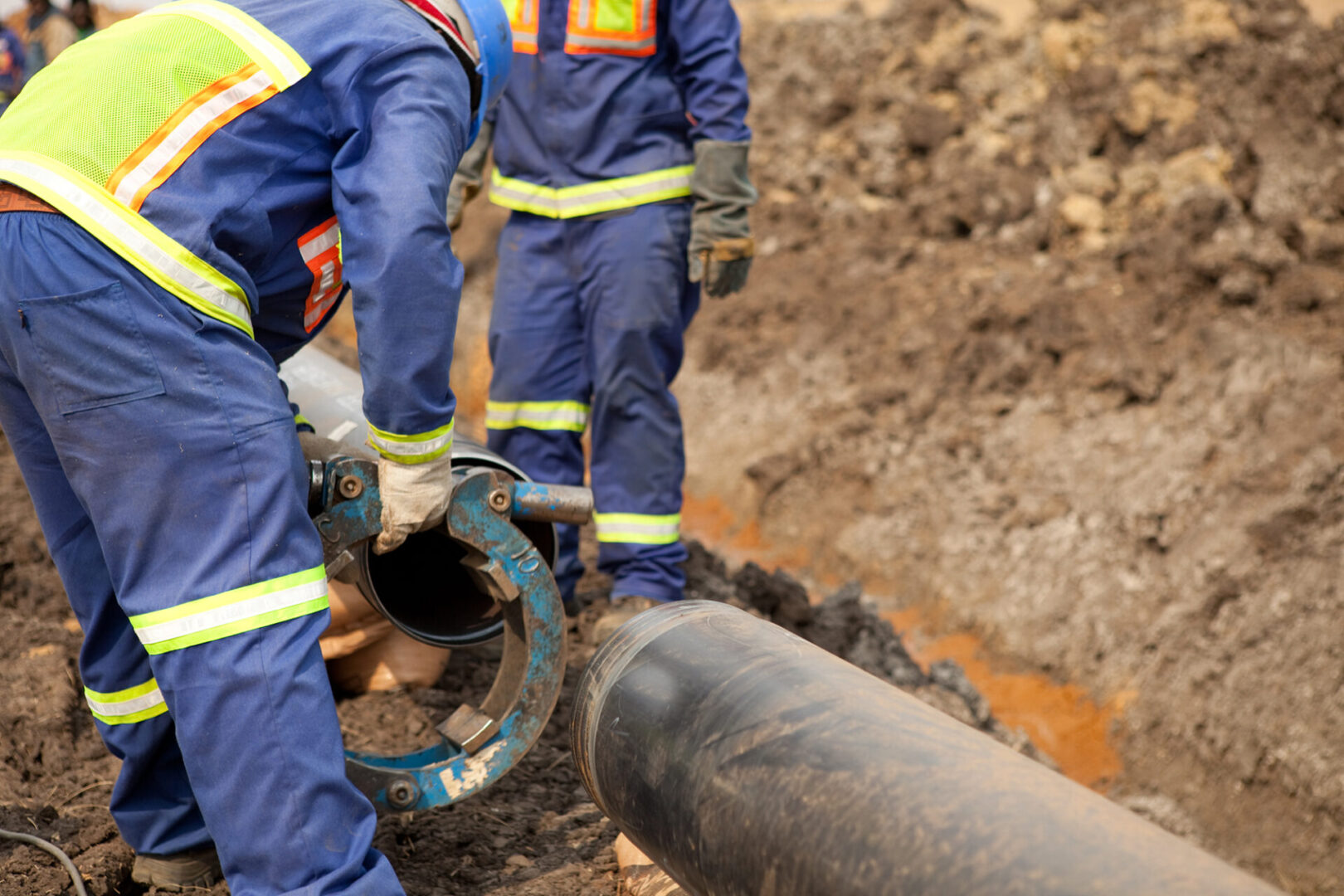 Male welder workers wearing protective high visibility clothing fixing and joining industrial construction oil and gas or water plumbing pipeline using an external pipe clamp outside on site